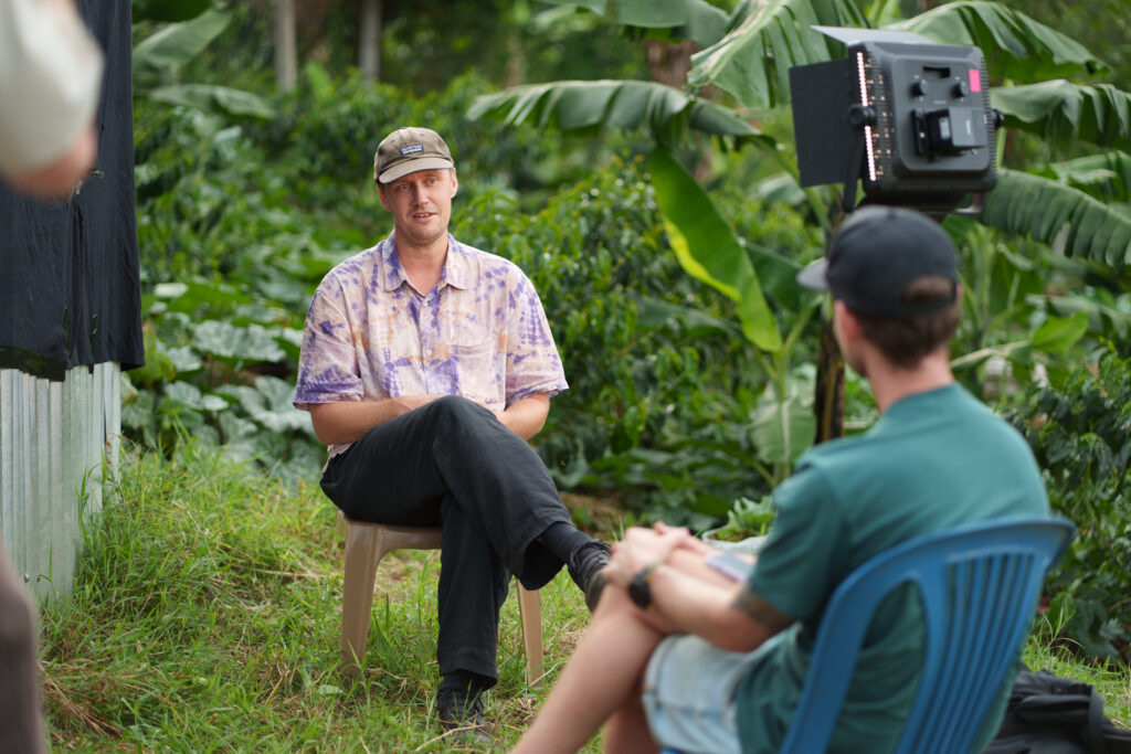 Lars Pilengrim sitting in a garden in Uganda, facing Robin Danehav and a camera during the interview.