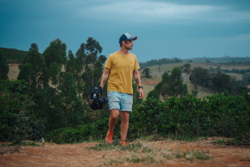 Robin Danehav in a yellow T-shirt walking through a coffee landscape in Brazil, camera in hand.