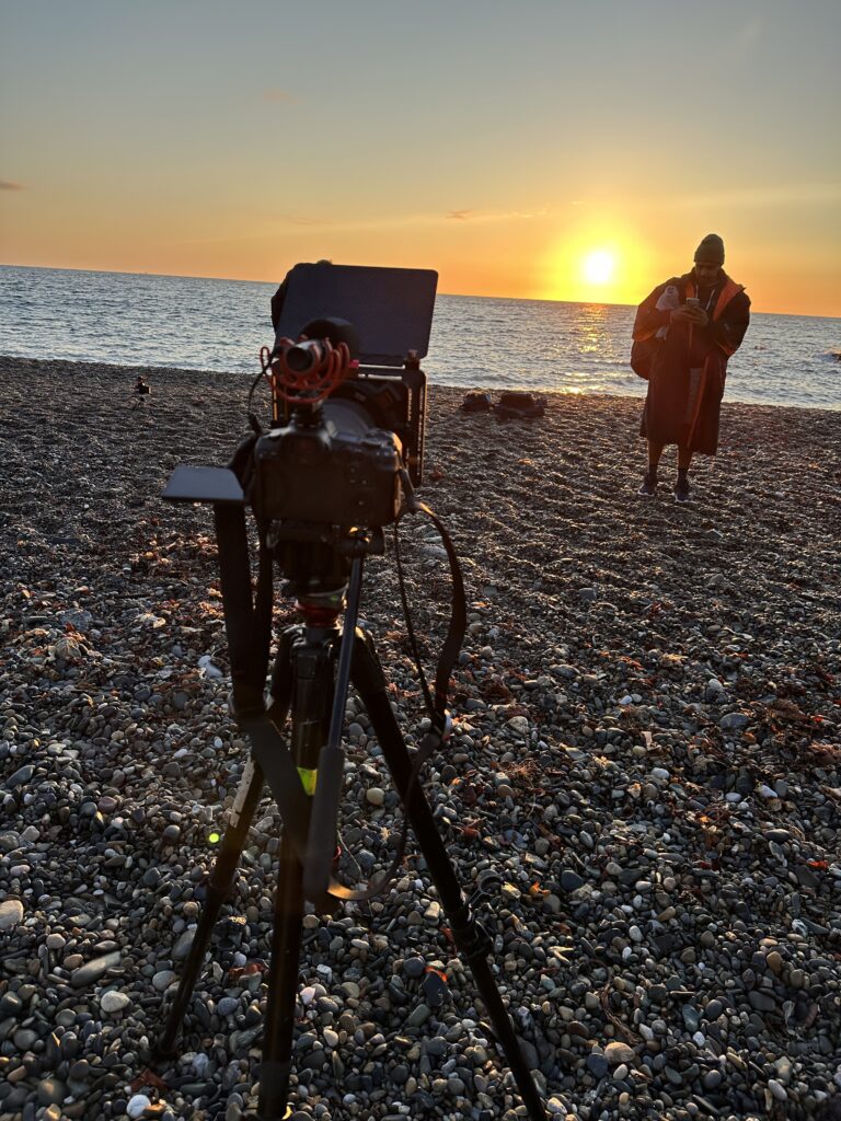 A camera on a tripod on a pebble beach at sunrise, with a person in a bathing robe at the water's edge.