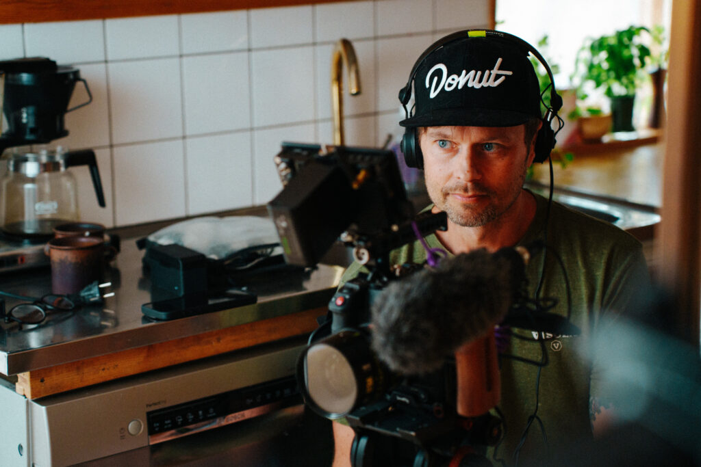 Robin Danehav in headphones and a cap behind a cinema camera with a microphone, filming in Oskar Kihlborg's kitchen.