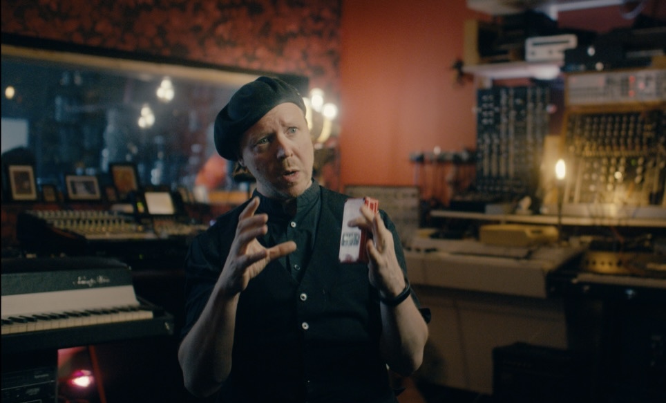 Mattias Olsson gesturing as he talks, surrounded by analog music gear in his studio.