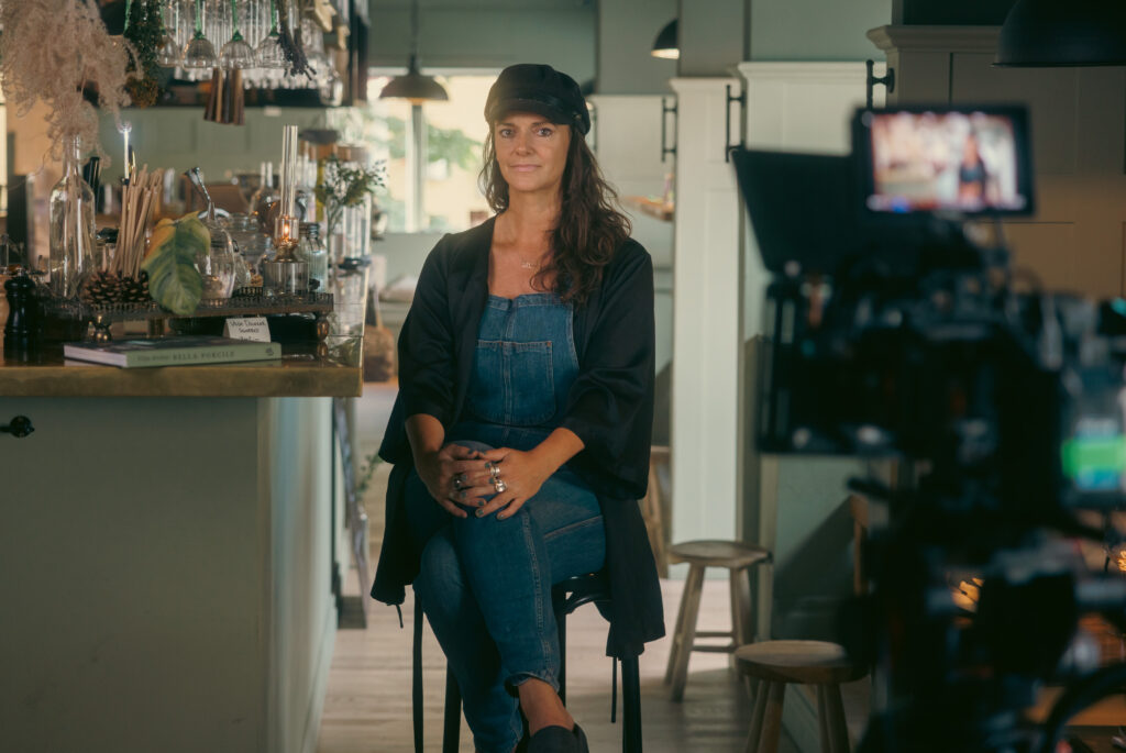 Bella Porcile sitting on a bar stool in a restaurant, with a camera and monitor set up for the interview.