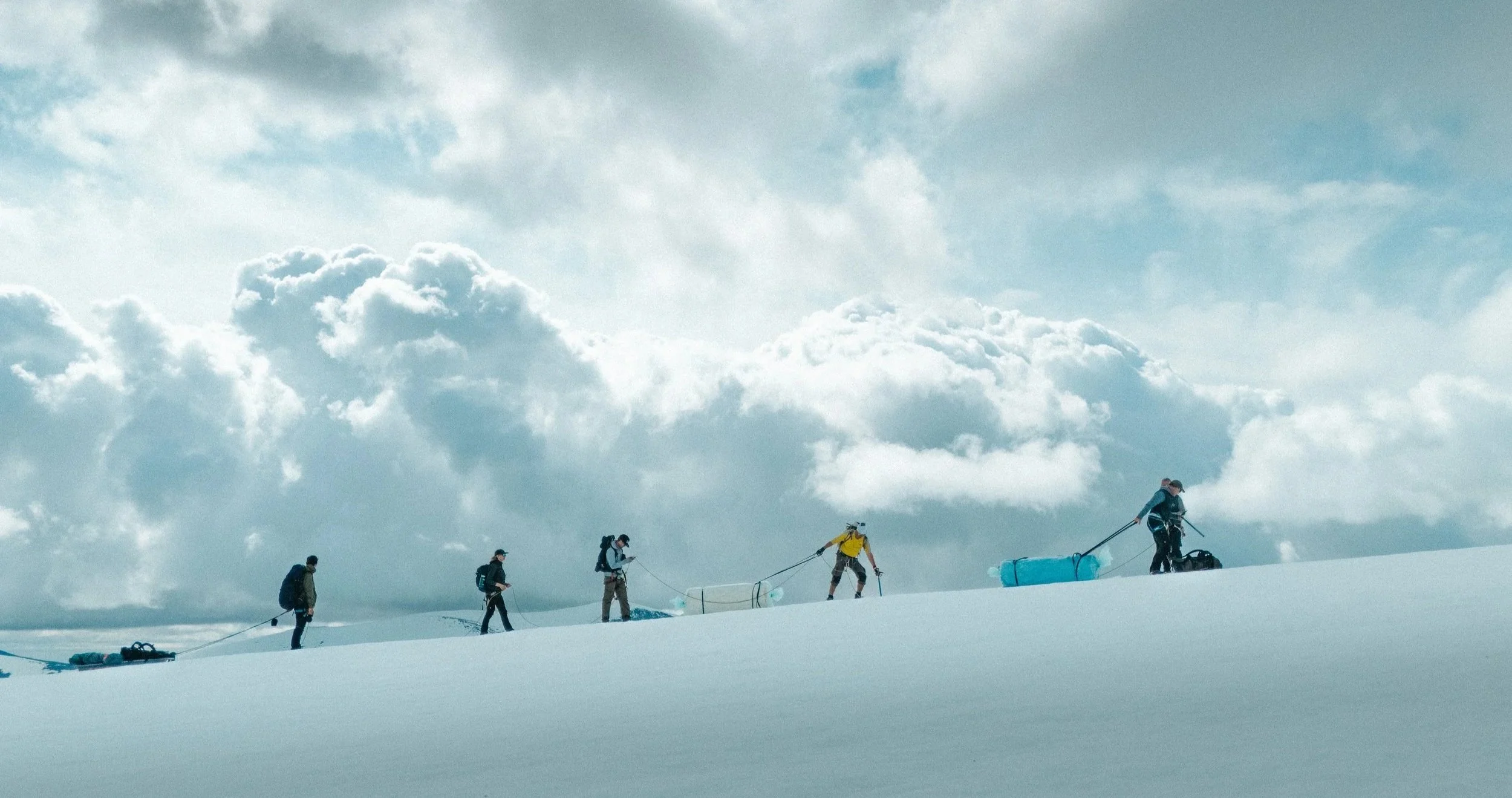 A line of people walking across the snow on Kebnekaise under a dramatic sky.