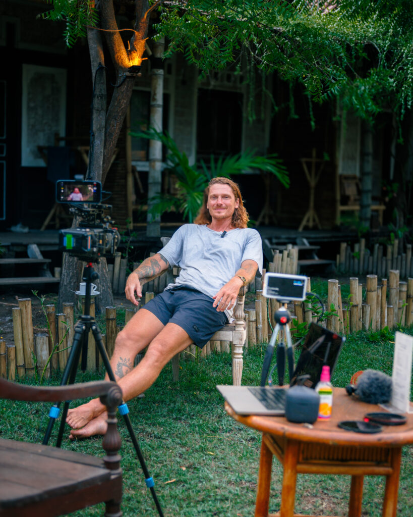 Martin Vrana sits in a garden chair facing two cameras on tripods, with a laptop and camera gear on a table beside him.