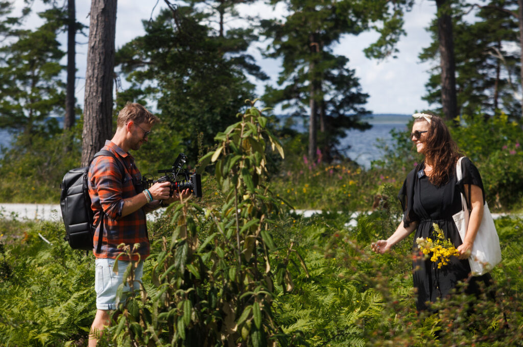 Robin Danehav filming as Bella Porcile picks plants in tall greenery by the sea on Gotland.
