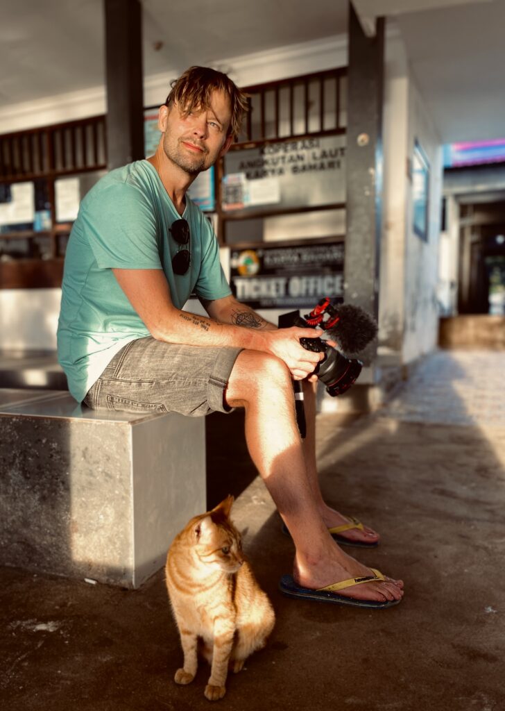 Robin Danehav sits on a ledge outside the ticket office at the Gili Trawangan harbour with a camera in his hands and a ginger cat at his feet.