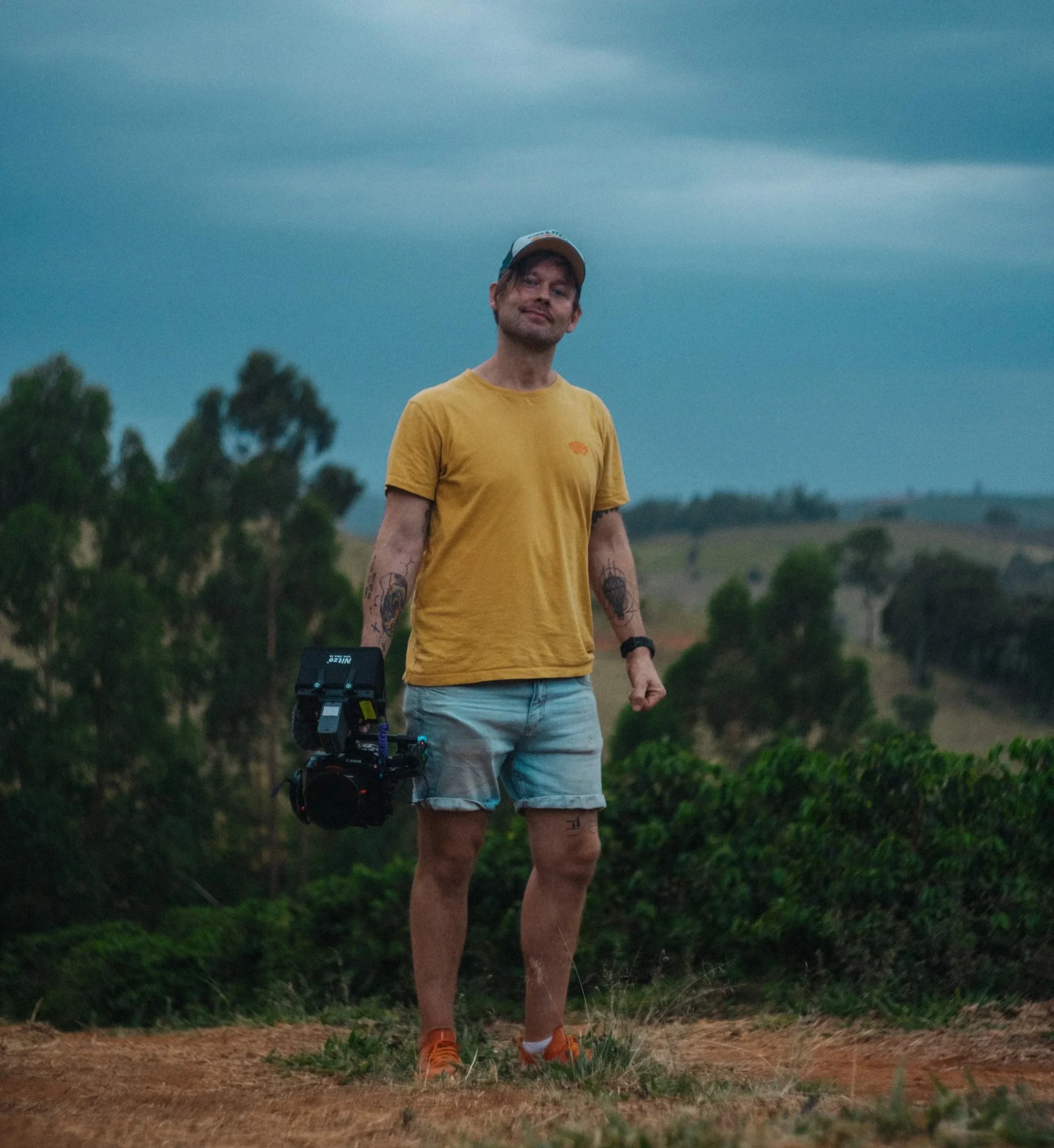 Robin Danehav in a yellow T-shirt, standing in a hilly landscape with his camera.