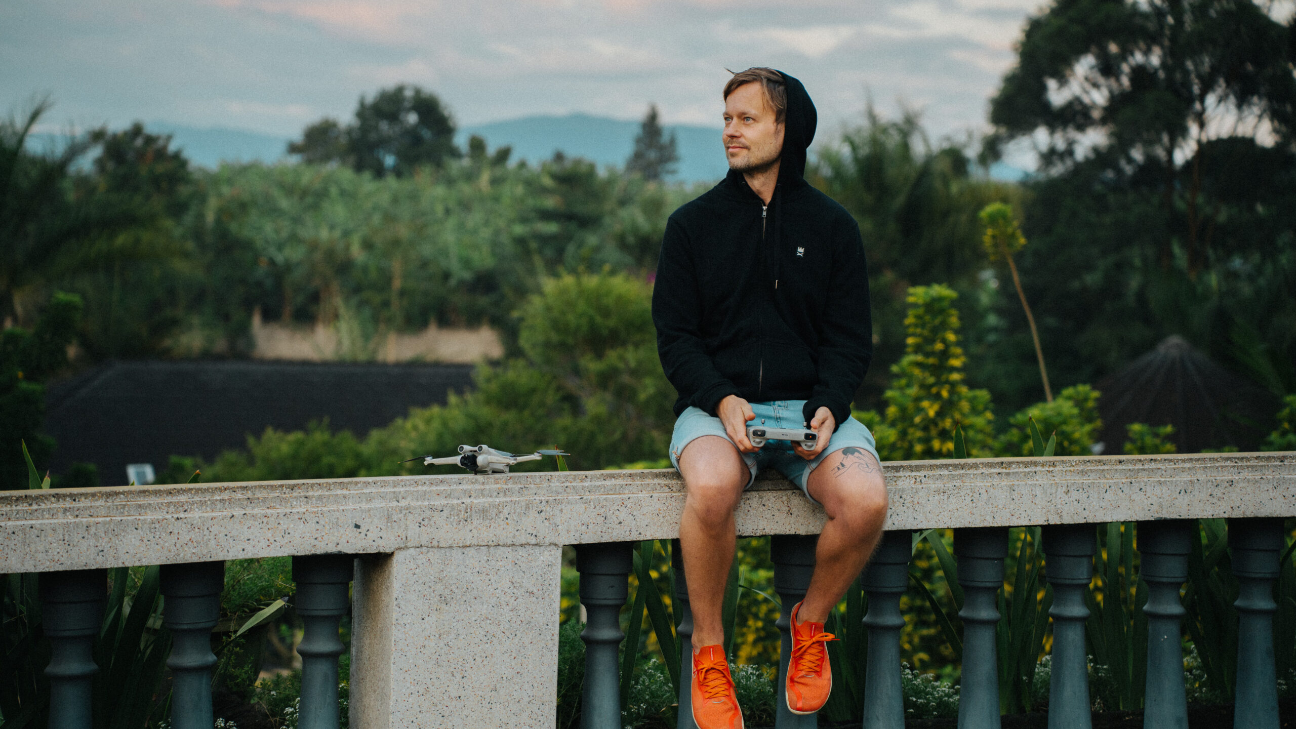Robin Danehav in a black hoodie and orange sneakers sitting on a stone railing with a drone controller, a small drone beside him and green hills behind.