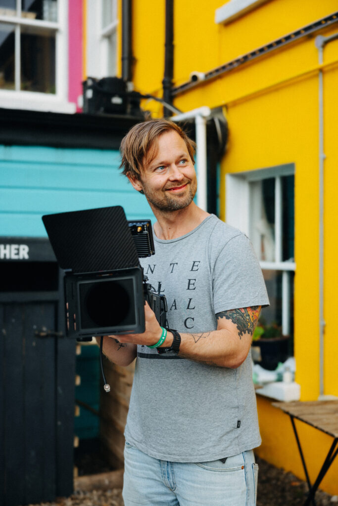 Robin Danehav holding a camera with a matte box outside a yellow café front in Greystones, Ireland.