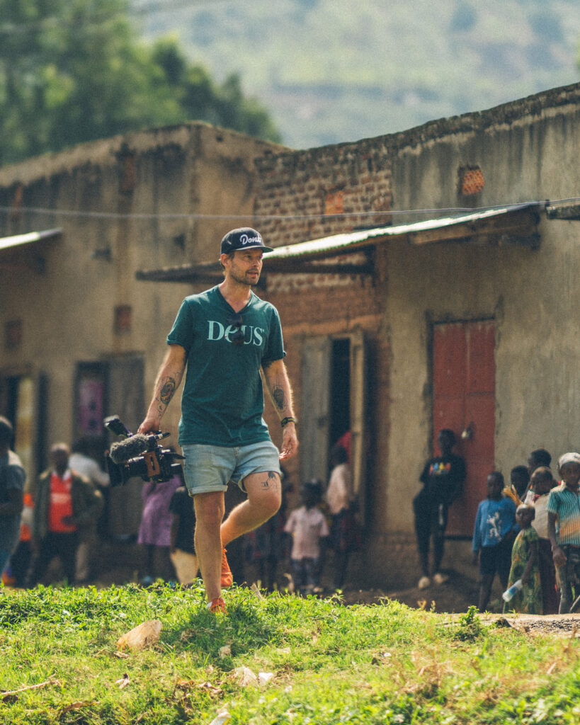 Robin Danehav walking across the grass in front of a village building in Uganda, with children in the background.